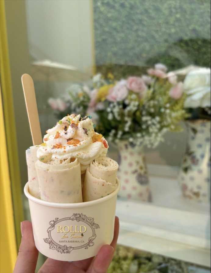 Fresh ROLLD ice cream rolls topped with whipped cream and colorful cereal, held in a branded cup beside flowers